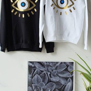 May include: Two hooded sweatshirts with a gold evil eye design. One sweatshirt is black and the other is white.