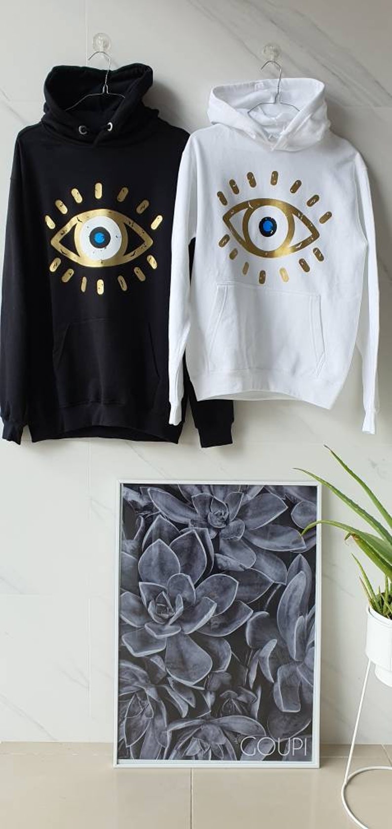 May include: Two hooded sweatshirts with a gold evil eye design. One sweatshirt is black and the other is white.