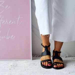Puede incluir: Un par de sandalias de cuero negro con correas de tobillo. Las sandalias tienen un dise&ntilde;o sencillo y las lleva una persona con pantalones blancos.