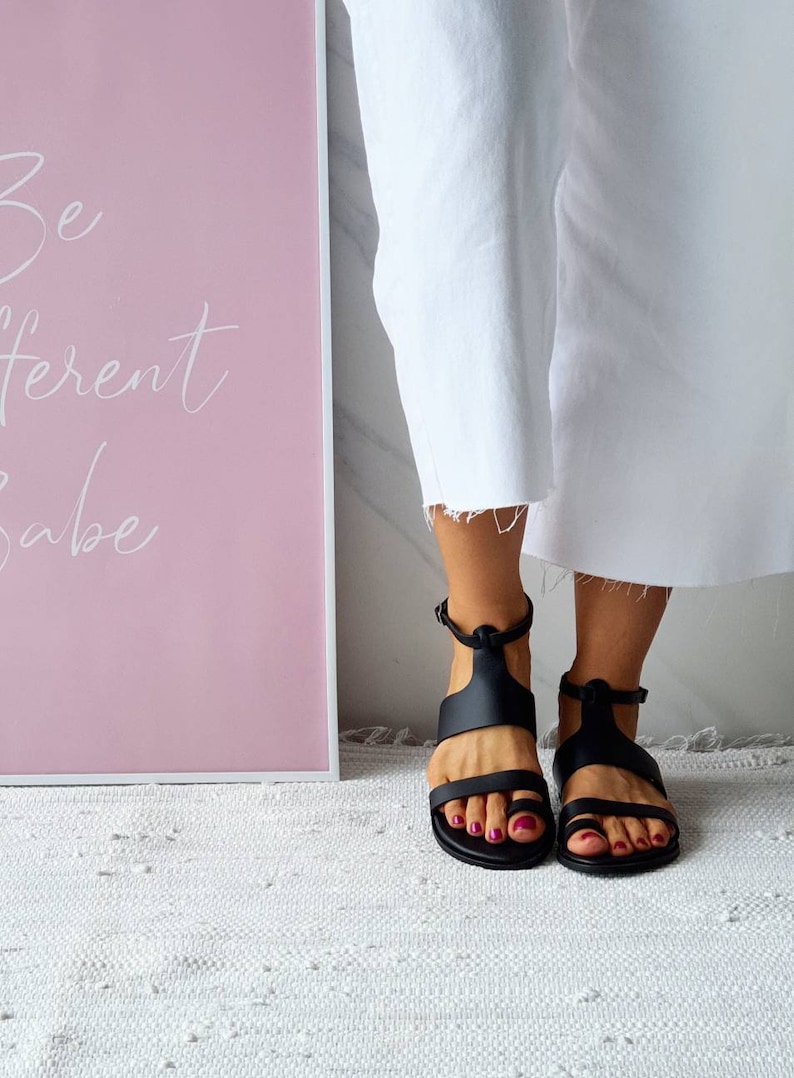 Puede incluir: Un par de sandalias de cuero negro con correas de tobillo. Las sandalias tienen un dise&ntilde;o sencillo y las lleva una persona con pantalones blancos.