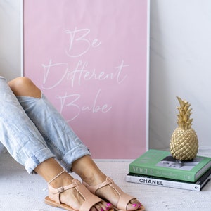 May include: A pair of light pink leather sandals with a t-strap design. The sandals are on a white rug with a pink framed poster that says "Be Different Babe" in white lettering.