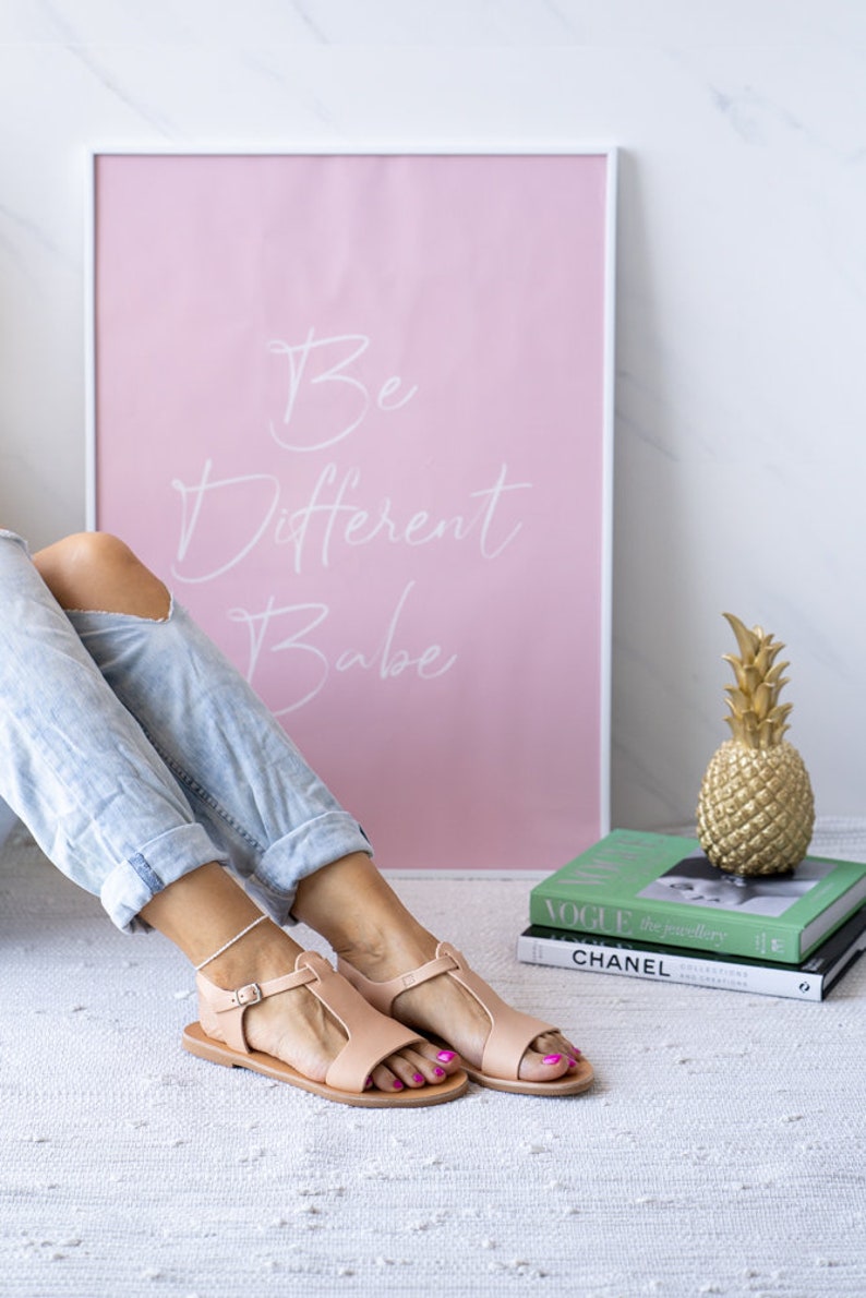 May include: A pair of light pink leather sandals with a t-strap design. The sandals are on a white rug with a pink framed poster that says "Be Different Babe" in white lettering.