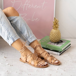 Puede incluir: Sandalias de cuero color canela con cordones que se atan en la pierna. Las sandalias tienen una suela plana y las lleva una persona con los pies descalzos. Las sandalias est&aacute;n sobre una alfombra blanca.