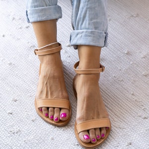 May include: A pair of tan leather sandals with ankle straps. The sandals have a simple design and are worn by a person with pink nail polish.