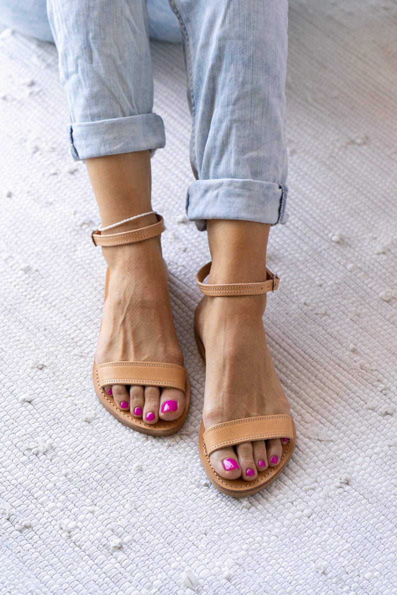 May include: A pair of tan leather sandals with ankle straps. The sandals have a simple design and are worn by a person with pink nail polish.