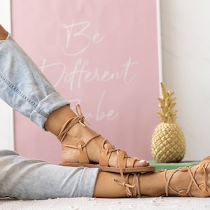 Puede incluir: Sandalias de cuero color canela con cordones que se atan en la pierna. Las sandalias tienen una suela plana y las lleva una persona con pantalones vaqueros azules.