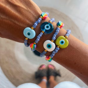 Può includere: Un set di quattro braccialetti con ciondoli a forma di occhio turco blu e bianco. Ogni braccialetto presenta un motivo di perline di colore diverso, tra cui arcobaleno, bianco e blu.