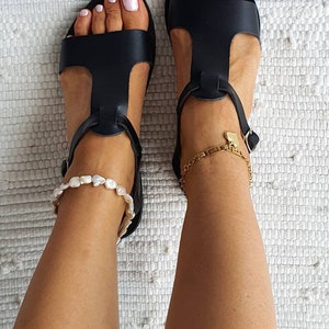 May include: A pair of black leather sandals with ankle straps. The sandals have a t-strap design and are worn on a person's feet. The person is wearing a light blue pair of jeans.