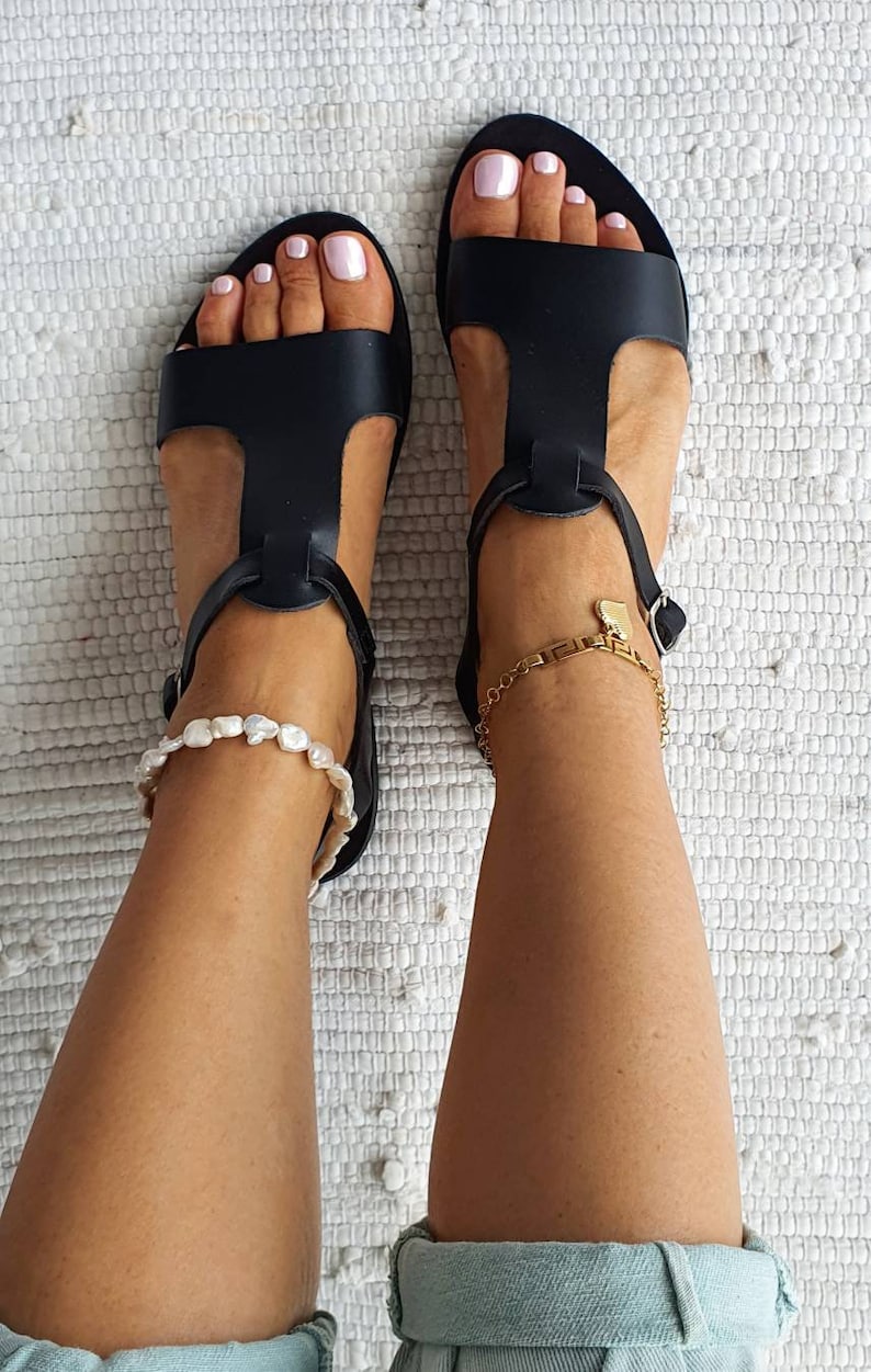 May include: A pair of black leather sandals with ankle straps. The sandals have a t-strap design and are worn on a person's feet. The person is wearing a light blue pair of jeans.