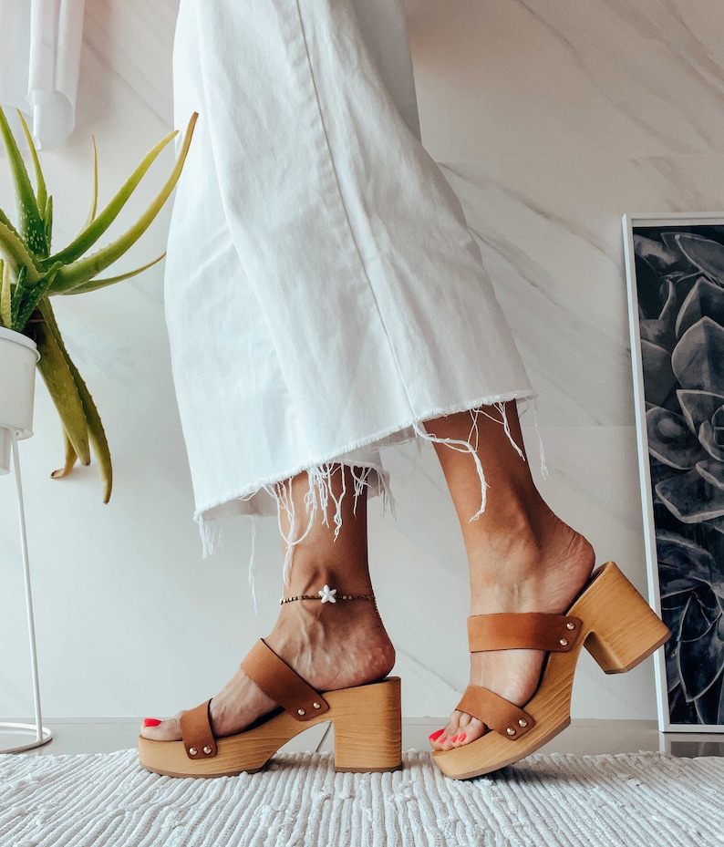 May include: A pair of brown leather platform clogs with a wooden heel. The clogs have a strap across the top of the foot and an ankle strap with a small star charm.