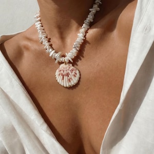 Puede incluir: Un collar de perlas blancas con un colgante de concha. El collar presenta perlas de forma irregular y un colgante de concha con borde dorado y patrones rosas y blancos. El collar se lleva sobre una camisa blanca.