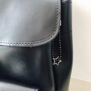 May include: Black leather backpack with a star-shaped zipper pull. The backpack has a flap with a zipper closure and a large pocket on the front.