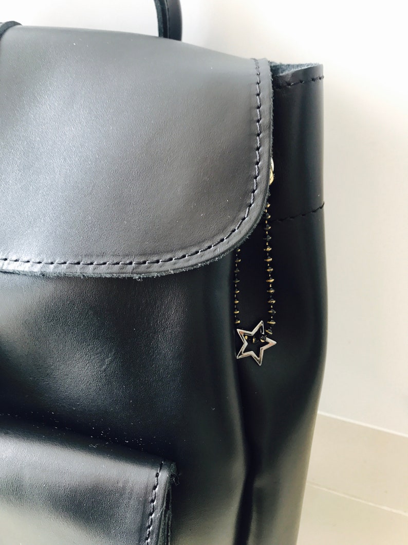 May include: Black leather backpack with a star-shaped zipper pull. The backpack has a flap with a zipper closure and a large pocket on the front.