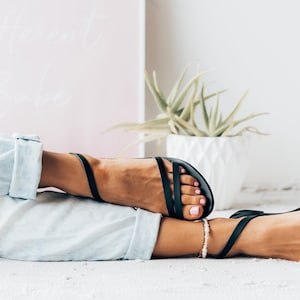 Puede incluir: Un par de sandalias de cuero negro con un dise&ntilde;o sencillo. Las sandalias tienen una tira fina que va alrededor del tobillo y una tira m&aacute;s gruesa que va a trav&eacute;s del pie. Las sandalias son usadas por una persona con los pies descalzos.