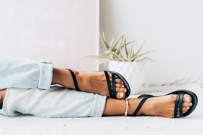 Puede incluir: Un par de sandalias de cuero negro con un dise&ntilde;o sencillo. Las sandalias tienen una tira fina que va alrededor del tobillo y una tira m&aacute;s gruesa que va a trav&eacute;s del pie. Las sandalias son usadas por una persona con los pies descalzos.