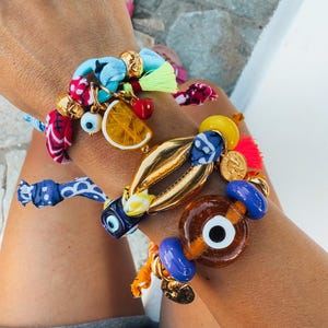 Könnte beinhalten: Eine Sammlung bunter Armbänder mit verschiedenen Anhängern, darunter eine Zitronenscheibe, Kirschen und eine augenförmige Perle. Die Armbänder haben goldfarbene Akzente, Stoffbänder und leuchtende Quasten. Das Thema ist verspielt und sommerlich.