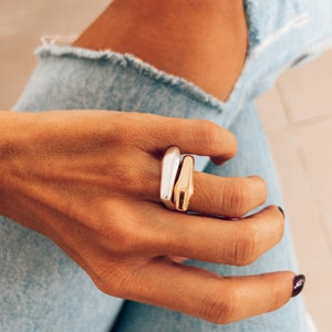 Può includere: Due anelli impilati su un dito. L'anello superiore è d'argento e l'anello inferiore è d'oro. Entrambi gli anelli hanno un design semplice e moderno.