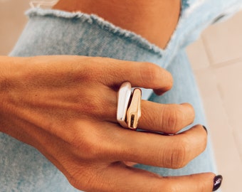 May include: Two rings stacked on a finger. The top ring is silver and the bottom ring is gold. Both rings are a simple, modern design.