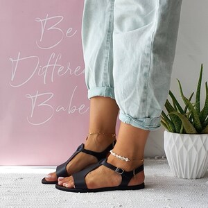 May include: Black leather sandals with a buckle closure and a t-strap design. The sandals are worn by a person with a light blue denim pants.