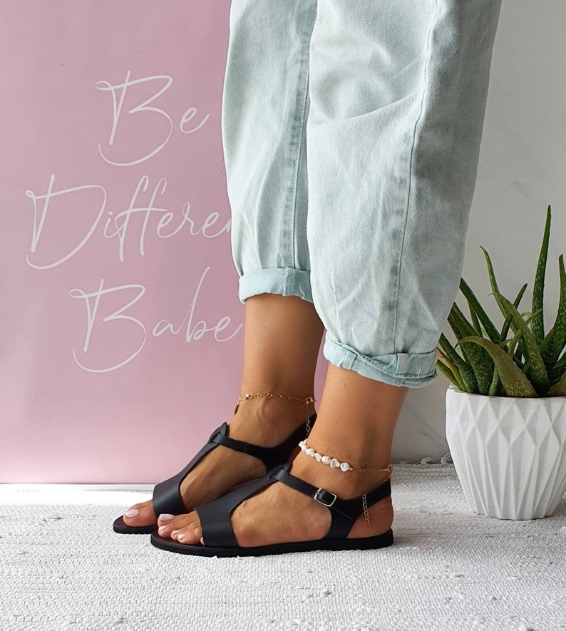 May include: Black leather sandals with a buckle closure and a t-strap design. The sandals are worn by a person with a light blue denim pants.