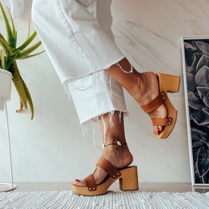 May include: A pair of brown leather platform sandals with a wooden heel. The sandals have two straps across the foot and an ankle strap. The model is wearing white pants and the sandals are on a white rug.