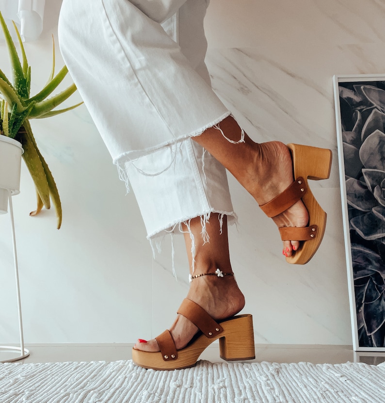 May include: A pair of brown leather platform sandals with a wooden heel. The sandals have two straps across the foot and an ankle strap. The model is wearing white pants and the sandals are on a white rug.