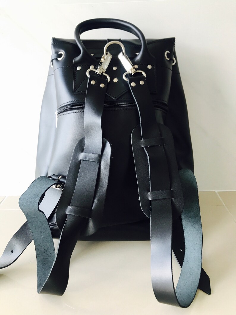 May include: Black leather backpack with silver hardware and adjustable straps. The backpack has a flap closure and a zippered pocket on the front.