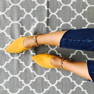 May include: A pair of yellow pointed-toe flats with gold chain ankle straps. The shoes are on a gray and white patterned rug.
