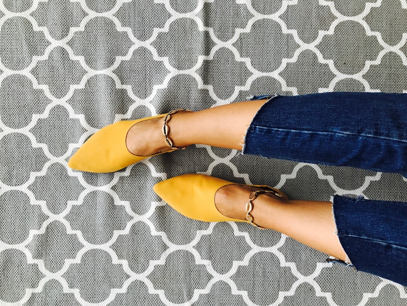 May include: A pair of yellow pointed-toe flats with gold chain ankle straps. The shoes are on a gray and white patterned rug.