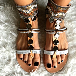 May include: A pair of silver and brown leather sandals with ankle straps decorated with sequins and beads. The sandals have a t-strap design and a round toe.