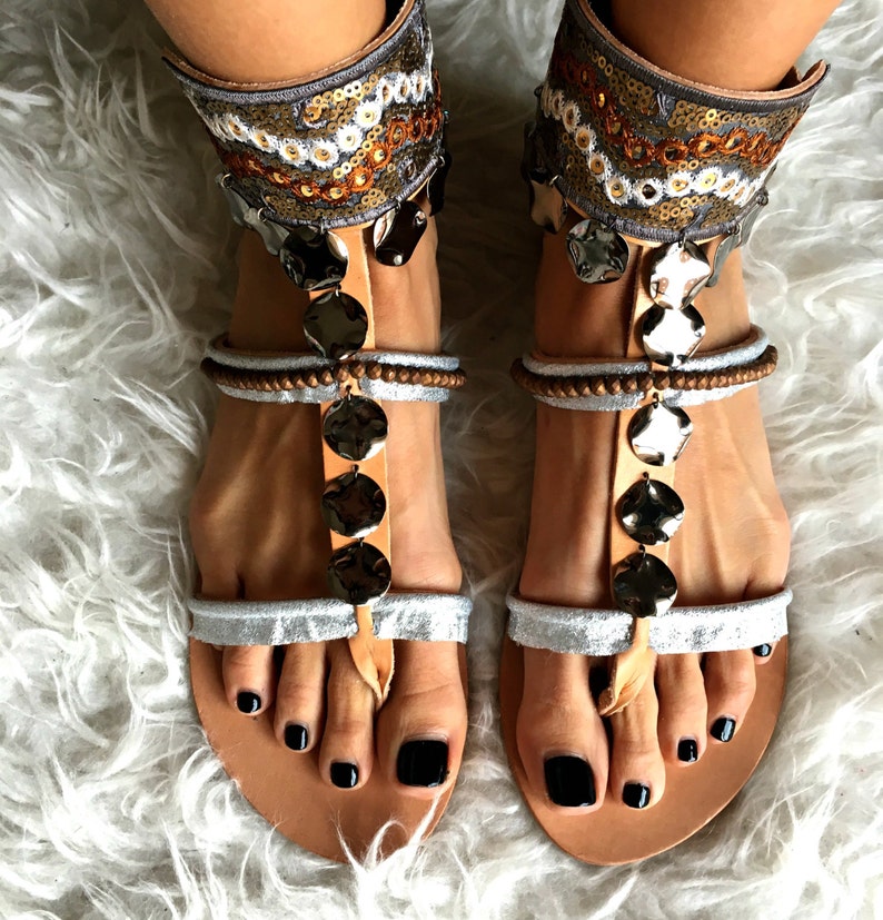 May include: A pair of silver and brown leather sandals with ankle straps decorated with sequins and beads. The sandals have a t-strap design and a round toe.