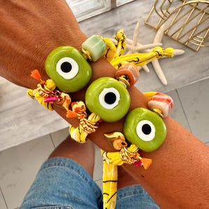 Könnte beinhalten: Armbänder in Augenform mit grünen Perlen und schwarzen Pupillen. Die Armbänder sind mit goldenen Ringen, orangefarbenen Quasten und gelbem Stoff verziert. Der Hintergrund umfasst ein goldenes Dekorationselement und Seesterne.
