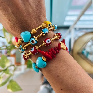 May include: A stack of bracelets with blue and red evil eye charms, turquoise beads, and coral beads. The bracelets are on a person's wrist.