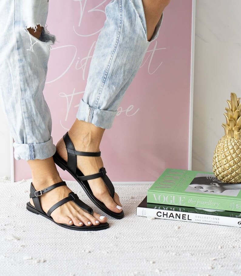 May include: A pair of black leather sandals with a buckle strap and a t-strap design. The sandals are worn by a person wearing blue jeans.