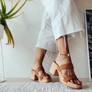 May include: A pair of brown leather platform sandals with a wooden heel. The sandals have two straps across the top of the foot and a buckle closure.