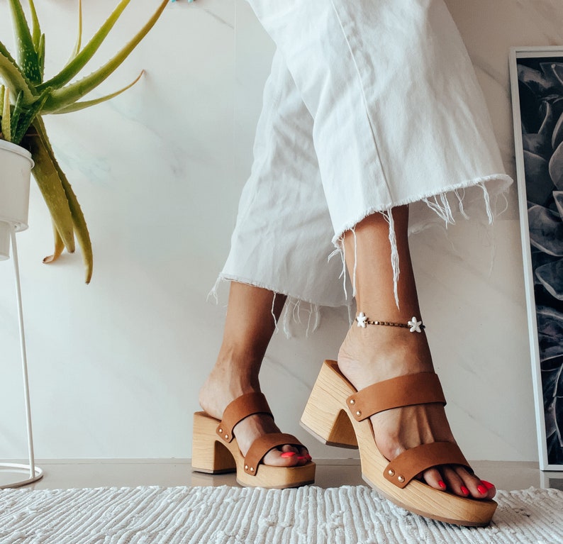 May include: A pair of brown leather platform sandals with a wooden heel. The sandals have two straps across the top of the foot and a buckle closure.