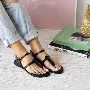 May include: A pair of black leather sandals with a buckle closure and a toe thong. The sandals are on a white rug with a stack of books in the background.