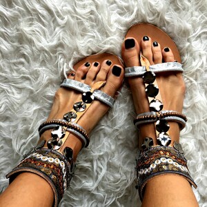 May include: Silver and brown sandals with black toe nails and beaded embellishments. The sandals have a strap that goes around the ankle and a buckle closure.