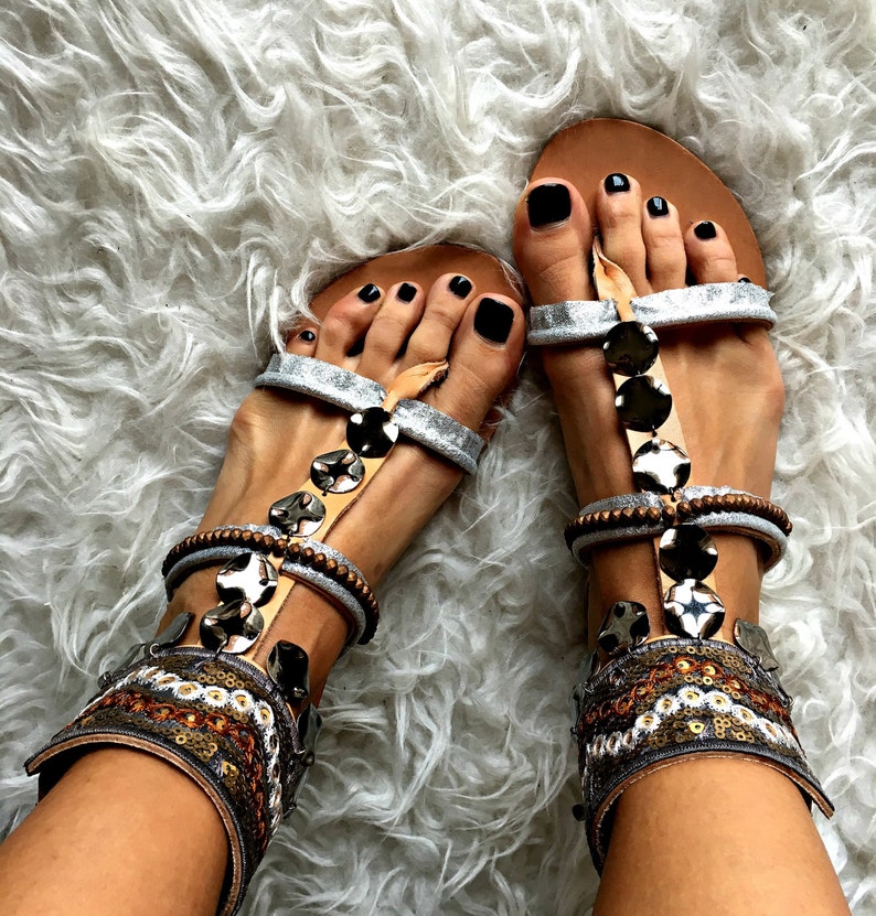 May include: Silver and brown sandals with black toe nails and beaded embellishments. The sandals have a strap that goes around the ankle and a buckle closure.