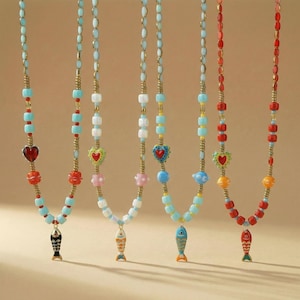 May include: A selection of beaded necklaces with vibrant colours. Each necklace has a different design, including heart, flower, and fish pendants. The beads are in shades of blue, red, white, and yellow. The necklaces are shown against a neutral backdrop.