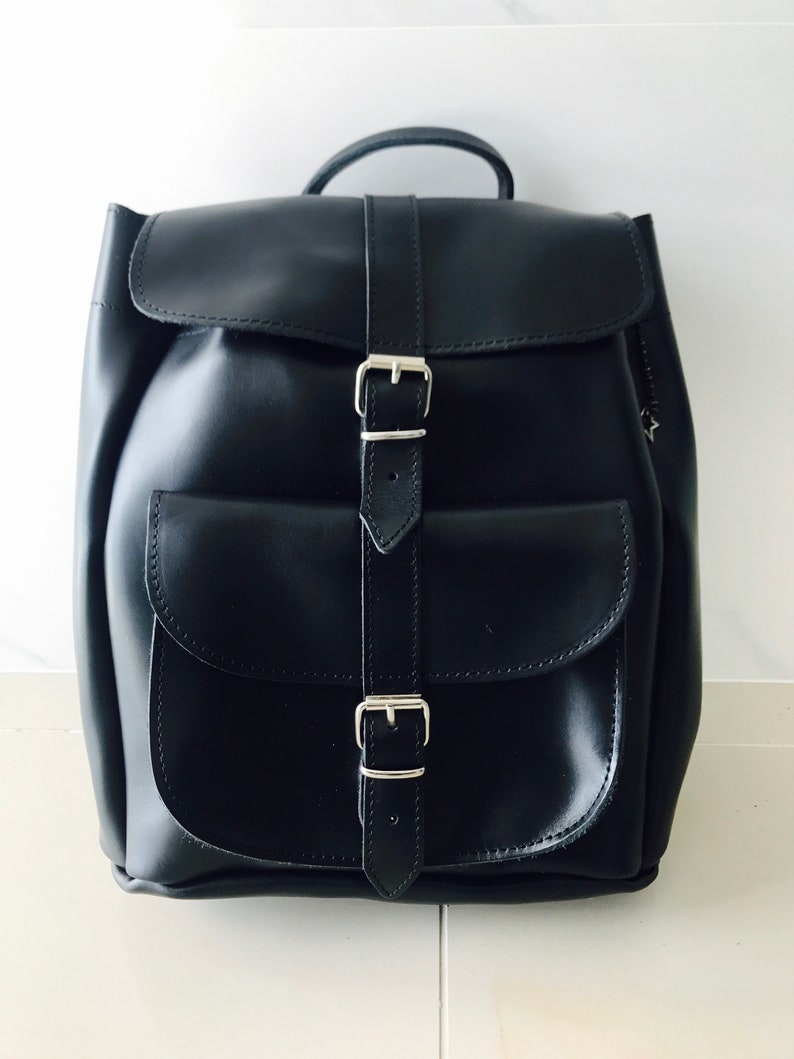 May include: A black leather backpack with two silver buckles and a flap closure. The backpack has a large pocket on the front.