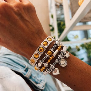 May include: A stack of bracelets with gold and silver beads and charms. The top bracelet is a gold and silver chain with square links and round gold accents. The charms include a star, a heart, and a coin with a profile image.