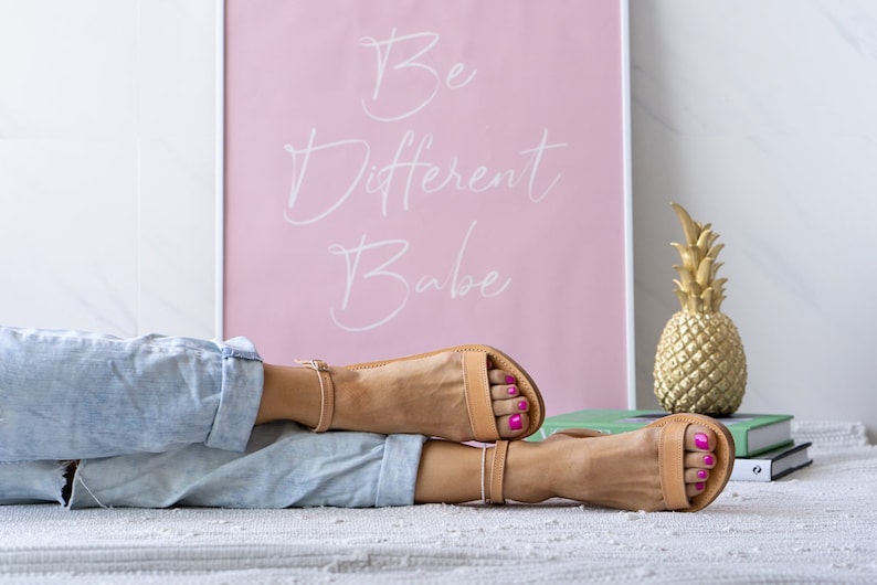 May include: A pair of tan leather sandals with ankle straps. The sandals are worn by a person lying on a white rug. A pink poster with the text "Be Different Babe" is in the background.