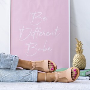 May include: A pair of light brown leather sandals with a T-strap design. The sandals have a light pink background with the text "Be Different Babe" written in white.