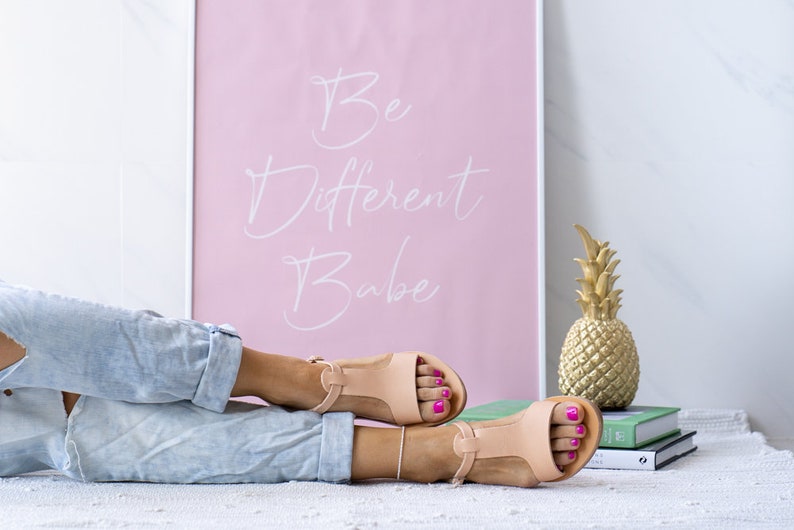 May include: A pair of light brown leather sandals with a T-strap design. The sandals have a light pink background with the text "Be Different Babe" written in white.