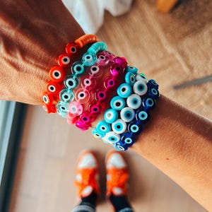 May include: A stack of colorful bracelets with evil eye charms. The bracelets are made of small, round beads in shades of blue, pink, orange, and turquoise. The evil eye charms are white with a blue center.