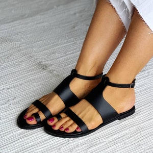 Puede incluir: Sandalias de cuero negro con dise&ntilde;o de tira en T y cierre de hebilla. Las sandalias tienen una suela plana y se llevan puestas.
