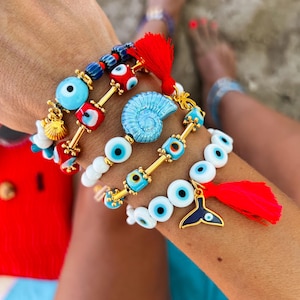 Può includere: Un set di braccialetti con ciondoli di occhio turco blu e bianco, perline rosse e blu e una catena dorata. I braccialetti includono un ciondolo a conchiglia blu e un ciondolo a coda di pesce blu con un occhio turco.