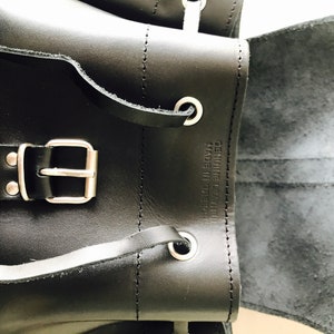 May include: Black leather backpack with a silver buckle and a flap that opens to reveal a black lining. The backpack has a label that reads "Genuine Leather Made in Greece."