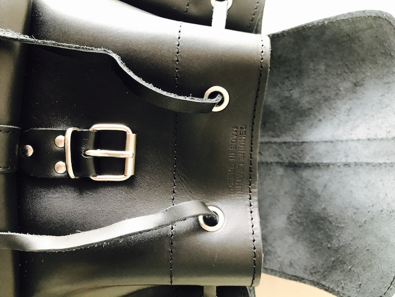 May include: Black leather backpack with a silver buckle and a flap that opens to reveal a black lining. The backpack has a label that reads "Genuine Leather Made in Greece."